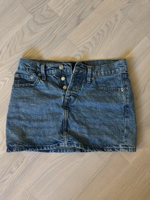 Blå jeanskjol med knappar - Säljer en klassisk blå jeanskjol från 157 med fyra silverfärgade knappar framtill. Kjolen är kort och har en rak passform, perfekt till sommaren. 