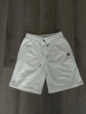 Vita sweatshorts från Moncler - Snygga vita sweatshorts från Moncler med elastisk midja och dragsko. Shortsen har två sidofickor och en liten Moncler-logga på vänster ben. Tillverkade i mjukt bomullsmaterial för en avslappnad och bekväm stil.