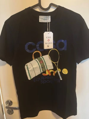 Svart Casablanca t-shirt med tennisprint - Casablanca svart t-shirt med färgglatt tryck av tennisväska, racket och boll på framsidan. Stor blå logga och text. Klassisk rund hals och korta ärmar. Perfekt för dig som gillar sportig stil och snygga detaljer.