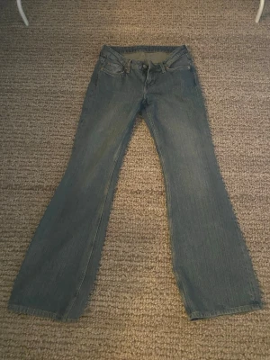 Weekday Nova Low Slim Bootcut Jeans - Säljer ett par Weekday Nova Low Slim Bootcut Jeans i klassisk blå denim. Jeansen har låg midja, smal passform upptill och utsvängda ben. Tillverkade i bomull med snygga detaljer och femficksmodell. Perfekta för dig som gillar retrovibbar och trendig siluett.
