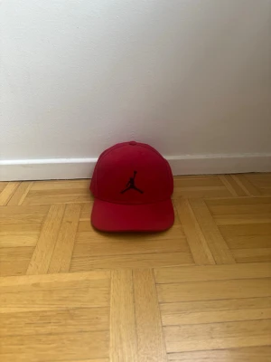 Röd Jordan keps med svart logga - Säljer en röd keps från Jordan med den klassiska svarta Jumpman-loggan framtill. Kepsen har böjd skärm och justerbar rem bak för perfekt passform. Tillverkad i ett mjukt och slitstarkt material som passar till streetwear och sportiga outfits.