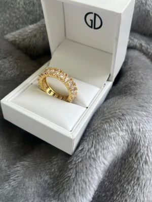 Guldfärgad ring med stenar från GD - Snygg guldfärgad ring från GULLDIA design med flera gnistrande, rektangulära stenar runt hela ringen. Ringen har en exklusiv känsla och levereras i en elegant vit ask. Perfekt för dig som vill sticka ut med ett statement-smycke. Ringen har storlek 19mm