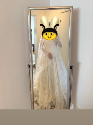 Vit lång spetsklänning med slöja - Elegant vit långklänning i spets med långärmade ärmar och vacker slöja. Klänningen har en klassisk, romantisk look med hög hals och utsvängd kjol. Perfekt för speciella tillfällen där du vill känna dig extra fin.