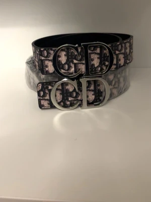 CD - Snyggt bälte med ikoniskt CD-spänne i silverfärg. Bältet har ett svart och ljust mönster med logotypdetaljer som ger en exklusiv vibe. Perfekt accessoar för att lyfta din outfit och visa din stil.