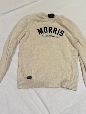 Beige stickad tröja Morris - Beige stickad tröja från Morris med svart text 'MORRIS COMPANY' på bröstet. Tröjan har rund halsringning, ribbade muddar och nederkant samt en liten logolapp nedtill. Perfekt för en avslappnad och stilren look.