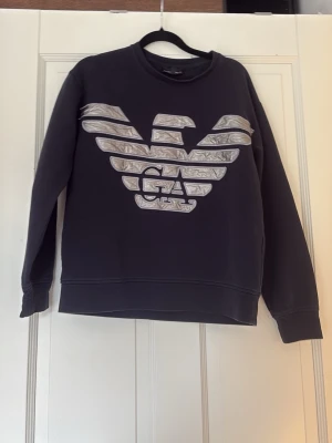 Svart sweatshirt från Emporio Armani - Cool svart sweatshirt från Emporio Armani med stort silverfärgat logotryck på bröstet. Tröjan har rund halsringning och är tillverkad i mjukt bomullsmaterial. Perfekt för dig som vill ha en stilren och trendig look.