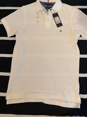 Vit pikétröja Tommy Hilfiger XS - Klassisk vit pikétröja från Tommy Hilfiger i slim fit-modell. Tröjan har korta ärmar, krage med knappar och den ikoniska lilla loggan broderad på bröstet. Tillverkad i bomull för en skön och luftig känsla.