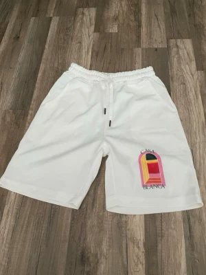 Vita shorts från Casablanca med tryck - Säljer ett par vita shorts från Casablanca med färgglatt tryck och logga på ena benet. Shortsen har elastisk midja med snörning och sidofickor. Perfekta för sommaren och riktigt sköna att bära till vardags eller när du chillar.