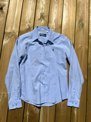 Ljusblå skjorta från Polo Ralph Lauren - Klassisk ljusblå skjorta från Polo Ralph Lauren med broderad logga på bröstet. Skjortan har lång ärm, knappar framtill och en stilren krage. Perfekt för en clean och snygg look. Storleken är XL i barnstorlek, så jag skulle säga att den är XS-S.