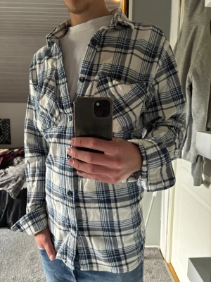 Rutig skjorta - Säljer en vit och blå rutig skjorta med klassisk krage och knäppning framtill. Skjortan har lång ärm och en bröstficka. Perfekt för en avslappnad och stilren look.
