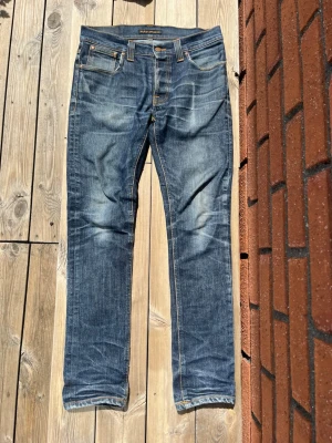 Blå Nudie Jeans med slitningar - Säljer ett par blå jeans från Nudie Jeans med klassisk femficksdesign och snygga slitningar. Jeansen har raka ben och kontrastsömmar, samt Nudie Jeans ikoniska broderi på bakfickorna. Materialet är denim i bomull.