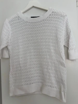 Vit stickad t-shirt med hålmönster - Säljer en vit stickad t-shirt med korta ärmar och snyggt hålmönster. Tröjan har ribbade muddar vid ärmslut och nederkant samt rund halsringning. Perfekt för lager på lager och ger en fräsch look till din outfit.
