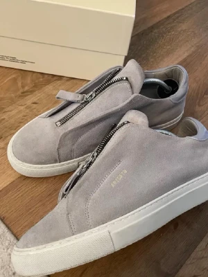 Gråa Axel Arigato sneakers med dragkedja - Snygga gråa sneakers från Axel Arigato i mocka med cool dragkedja framtill istället för klassiska snören. Skorna har vit sula och minimalistisk design med diskret logga på sidan. Perfekt för dig som gillar stilrena och moderna skor. Inget box medföljer 