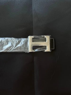 Svart skärp med silverfärgat H-spänne - Snyggt svart bälte med ett stort, silverfärgat H-format spänne och vita sömmar längs kanterna. Bältet har en stilren och modern look, perfekt för att lyfta vilken outfit som helst. Tillverkat i skinn med detaljer i metall.