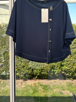 Mörkblå kortärmad blus från Zara - Stilren mörkblå blus från Zara med korta ärmar och dekorativa silverfärgade knappar framtill. Blusen har vita kontrastsömmar som ger en cool touch och är tillverkad i ett mjukt, lätt material. Perfekt att matcha med jeans eller kjol för en trendig look.