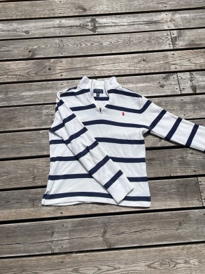 Randig stickad tröja Polo Ralph Lauren - Vit stickad tröja med mörkblå horisontella ränder från Polo Ralph Lauren. Tröjan har halv dragkedja vid halsen och röd broderad logga på bröstet. Långärmad och i mjukt material, perfekt för en avslappnad och stilren look.