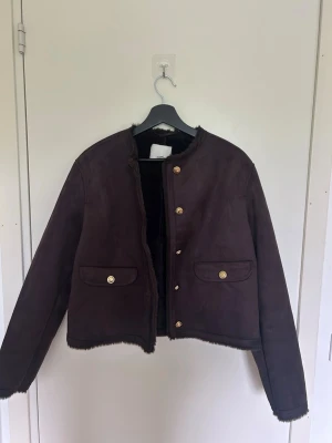 Mörkbrun pälsjacka från H&M - Snygg mörkbrun pälsjacka från H&M med guldfärgade knappar och två fickor framtill. Jackan har en rak, boxig passform och mjukt foder i fuskpäls som håller dig varm under kalla dagar. Perfekt för dig som vill ha en trendig och mysig vinterjacka.