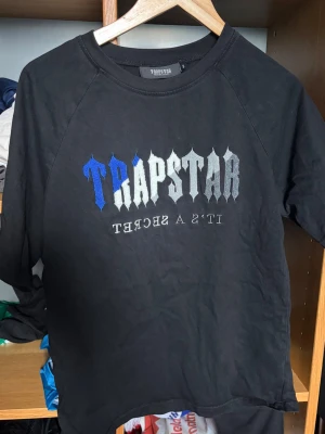 Svart Trapstar hoodie med tryck - Svart hoodie från Trapstar med stort broderat TRAPSTAR-tryck i blått och grått på bröstet samt texten 'IT'S A SECRET' under. Klassisk passform med rund hals och långa ärmar. Perfekt för dig som gillar streetwear och vill sticka ut.
