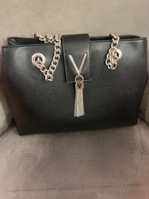 Svart handväska från Zalando i märket Valentino  - Snygg svart handväska från Valentino med kedjehandtag i silverfärg och dekorativt V-hänge med tofs framtill. Väskan är tillverkad i strukturerat konstläder och har en stilren, rektangulär form. Perfekt accessoar för att lyfta din outfit.