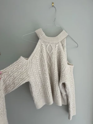 Beige stickad tröja med öppna axlar - Trendig beige stickad tröja med öppna axlar och bred ribbad krage. Tröjan har ett snyggt flätmönster framtill och långa ärmar. Perfekt för dig som vill ha en unik och stilren look. Materialet är mjukt och känns lyxigt.