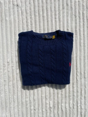 Mörkblå kabelstickad tröja Polo Ralph Lauren - Snygg mörkblå kabelstickad tröja från Polo Ralph Lauren med klassisk rund halsringning och röd broderad logga på bröstet. Tröjan har långa ärmar och ribbade muddar vid ärmslut och nederkant. Perfekt för dig som gillar stilrena och tidlösa plagg.