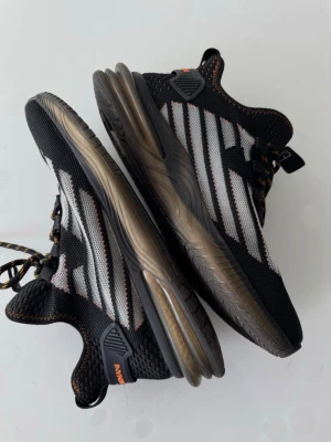 Svarta sneakers från Armani - Snygga svarta sneakers från Armani med vita och grå detaljer samt orange sömmar. Skorna har en sportig design med meshmaterial för bra andningsförmåga, tjock sula i brunt och svarta skosnören med orange inslag. Perfekta för en trendig streetwear-look.