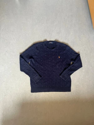 Mörkblå kabelstickad tröja Polo Ralph Lauren -  En mörkblå stickad tröja från Polo Ralph Lauren med kabelstickat mönster. Ralph lauren loggan broderad i orange på bröstet. Perfekt för höst/vår. Tröjan är i storlek L. Skriv ifall ni har frågor så svarar jag så snabbt som möjligt, skickar inom 1-2 dagar max så länge jag är i Sverige!👍