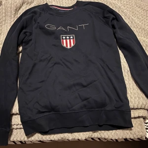 Mörkblå sweatshirt från GANT - Mörkblå sweatshirt från GANT med broderad logga och ett sköldmärke med stjärnor och ränder på bröstet. Tröjan har rund halsringning, långa ärmar och ribbade muddar vid ärmslut och nederkant. Klassisk och stilren design.