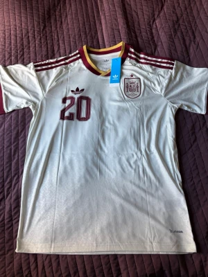 Vit Spanien fotbollströja Adidas - Säljer en vit Spanien fotbollströja från Adidas med vinröda detaljer, nummer 20 och namnet Pedri på ryggen. Tröjan har korta ärmar, rund hals med gul och vinröd kant samt broderad landslagssköld på bröstet. Perfekt för dig som älskar fotboll och vill stötta Spanien.