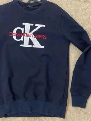 Mörkblå Calvin Klein sweatshirt - Mörkblå sweatshirt från Calvin Klein Jeans med stort vitt CK-tryck och röd logga framtill. Klassisk rund halsringning och ribbade muddar vid ärmslut och nederkant. Perfekt för en avslappnad och stilren look.