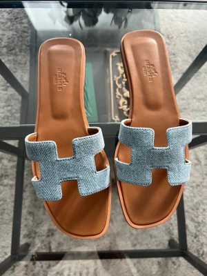 Sommar sandaler i denim och läder - Säljer ett par sandaler med klassisk H-design. Ovandelen är i ljusblå denim och sulan i brunt läder. Sandalerna har öppen tå och fyrkantig form, vilket ger en trendig och avslappnad look. Perfekta för dig som gillar exklusiva detaljer och stilren design. Passar till str 40