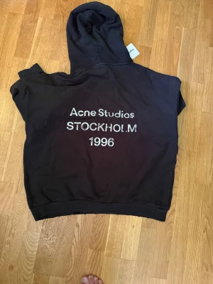 Svart hoodie Acne Studios Stockholm - Svart hoodie från Acne Studios med stor huva och tryck på ryggen: 'Acne Studios STOCKHOLM 1996'. Mjuk bomullskvalitet och ribbade muddar. Perfekt för dig som gillar streetwear och vill ha något stilrent med coolt tryck.