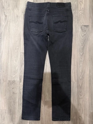 Nudie Jeans Lean Dean Jeans Mörkgrå W30/L32 - Slim Tapered Fit - Nudie Lean Dean mörkgrå jeans med slim tapered fit passform som sitter smalt men bekvämt och funkar till allt, storlek W30/L32, midja 39,5 cm, ytterbenslängd 102 cm, benöppning 15,5 cm, modellen är 185cm & väger 65kg, i fint skick. Litet hål i skrevet.