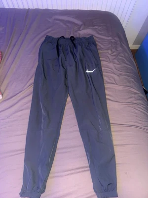 Nike svart träningsset med huva - Säljer ett svart träningsset från Nike med byxor och matchande jacka med huva. Båda plaggen har den klassiska vita Nike-loggan och är tillverkade i lätt, snabbtorkande material. Jackan har dragkedja och fickor, byxorna har resår i midjan och benslut.