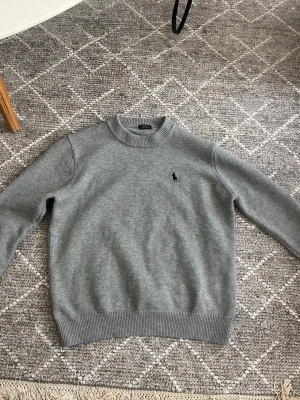 Grå stickad tröja från Polo Ralph Lauren - Klassisk grå stickad tröja från Polo Ralph Lauren med rund halsringning och ribbade muddar. Tröjan har den ikoniska lilla broderade loggan på bröstet. Perfekt basplagg i mjukt material som funkar året runt.