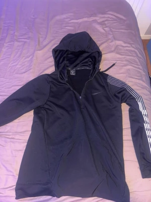 Svart Nike hoodie med half zip - Svart hoodie från Nike med huva och half zip-dragkedja. Snygga vita ränder längs ena ärmen och diskret Nike-logga på bröstet. Tillverkad i mjukt och sportigt syntetmaterial, perfekt för en avslappnad och sportig stil.