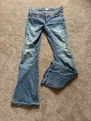 Blå bootcut jeans med slitningar - Säljer ett par ljusblå bootcut jeans med slitningar på framsidan och broderade detaljer på bakfickorna. Jeansen har låg midja och klassisk femficksdesign. använda ca 2 gånger, upp sprettade där nere. Längden passar bra för mig som är 172