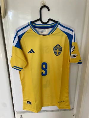 Svensk landslagströja Adidas gul/blå - Säljer en officiell svensk landslagströja från Adidas i gult och blått med nummer 9 på bröstet. Tröjan har korta ärmar, broderad landslagssköld och Adidas-logga. Tillverkad i lätt och ventilerande material, perfekt för fotboll eller träning.