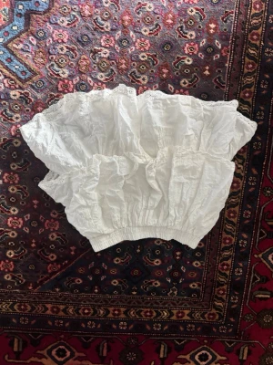 Vit volangkjol med spetsdetaljer - Söt vit kortkjol med flera volanger och spetsdetaljer längs kanterna. Kjolen har resår i midjan för en bekväm passform och är gjord i ett lätt bomullstyg. Perfekt för dig som gillar romantisk och luftig stil.