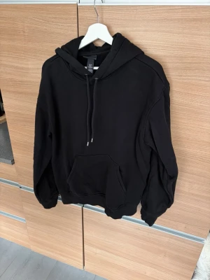 Svart hoodie från H&M med huva - Klassisk svart hoodie från H&M med stor känguruficka framtill och justerbar dragsko i huvan. Tillverkad i mjukt bomullsmaterial och har ribbade muddar vid ärmslut och nederkant. Perfekt för en avslappnad och stilren look.