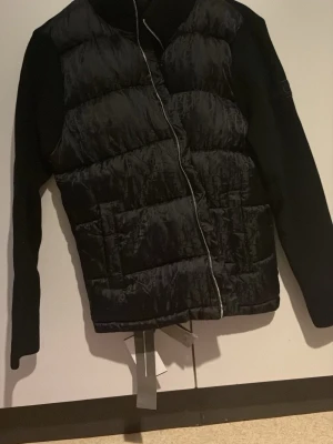Svart dunväst från Dior - Snygg svart dunväst från Dior med quiltad front och stickade ärmar. Västen har dragkedja framtill och justerbar nederkant med band. Klassisk Dior-logga på ärmen. Perfekt för lager-på-lager och streetwear-vibe.