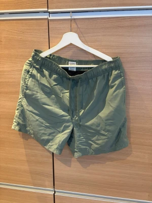 Olivgröna badshorts med dragsko - Säljer ett par olivgröna badshorts med elastisk midja och dragsko. Shortsen har en bakficka med lock och är tillverkade i ett snabbtorkande material, perfekta för stranden eller poolen. Klassisk och enkel design som funkar till det mesta.