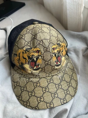 Gucci keps med tigertryck och mesh - Säljer en snygg Gucci keps med klassiskt GG-mönster i beige och brunt, två stora tigertryck framtill och svart mesh baktill. Justerbar rem med grönt och rött band bak. Säljs ej i butik längre, äkta!
