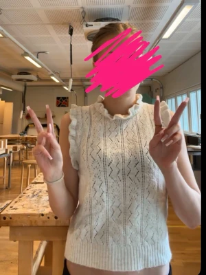 Vit stickad ärmlös topp med volang - Säljer en söt vit stickad topp med volangdetaljer vid axlarna och halsen. Toppen är ärmlös och har ett diskret hålmönster framtill som ger en trendig look. Perfekt att styla med jeans eller kjol för en fräsch outfit.