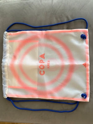 Vit och rosa gympåse från Adidas - Snygg gympåse från Adidas med texten COPA PURE III. Väskan är vit med rosa cirkelmönster och har blåa snören. Tillverkad i lätt syntetmaterial, perfekt för att bära träningskläder eller skor. Enkel och stilren design med dragskostängning.