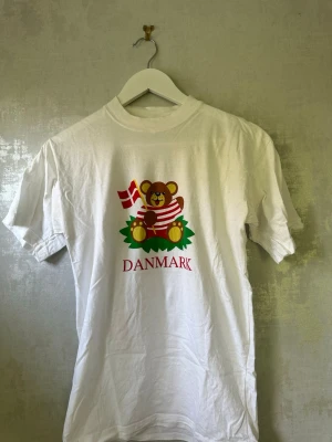 Vit Danmark t-shirt med björntryck - Vit t-shirt med ett gulligt tryck av en björn som håller en dansk flagga och texten 'DANMARK' under. T-shirten har rund hals och korta ärmar. Tillverkad i 100% bomull, perfekt för dig som gillar Danmark eller vill ha en söt souvenir.