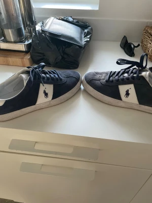 Mörkblåa sneakers från Polo Ralph Lauren - Snygga mörkblå sneakers från Polo Ralph Lauren med vit panel på sidan och klassisk logga. Skorna har låga skaft, snörning och ovandel i mocka. Perfekta för dig som gillar stilrena och sportiga skor med en ikonisk touch. Knappt använda och är i väldigt bra skick!