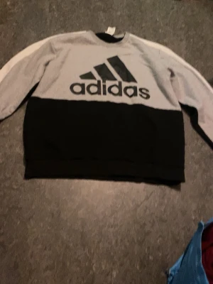 Grå och svart sweatshirt Adidas - Stickad tröja från Adidas i grått och svart med stor logga framtill. Tröjan har vita och svarta ränder längs ärmarna och rund halsringning. Perfekt för en sportig och avslappnad look.