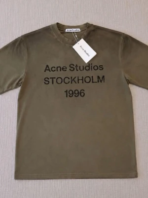 Olivgrön t-shirt Acne Studios - Olivgrön t-shirt från Acne Studios med svart text 'Acne Studios STOCKHOLM 1996' på bröstet. Klassisk rund hals och korta ärmar. Materialet är mjuk bomull och passformen är normal. Perfekt för dig som gillar stilrena och trendiga plagg.