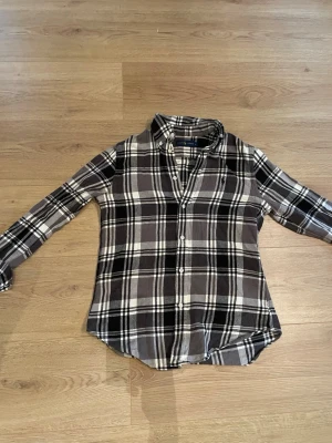 Rutig skjorta från Ralph Lauren - Säljer en klassisk rutig skjorta från Ralph Lauren i svart, grått och vitt. Skjortan har långa ärmar, knappar framtill och en liten broderad logga på bröstet. Perfekt för en avslappnad men snygg look.